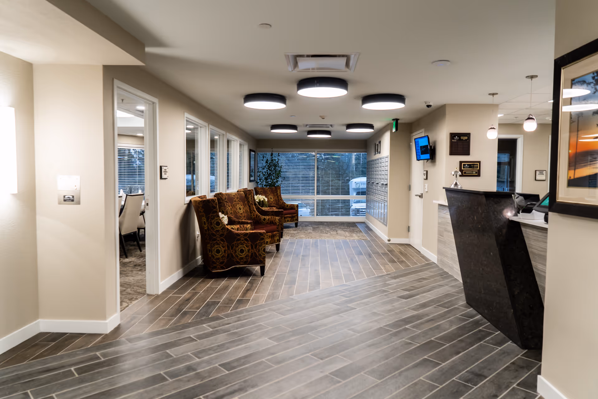 Interior view of a senior living facility lobby area with a modern reception desk on the right, three patterned armchairs along the left wall, large windows at the back, and a hallway leading to other rooms. The floor has a wood-like tile design, and the ceiling features round light fixtures.