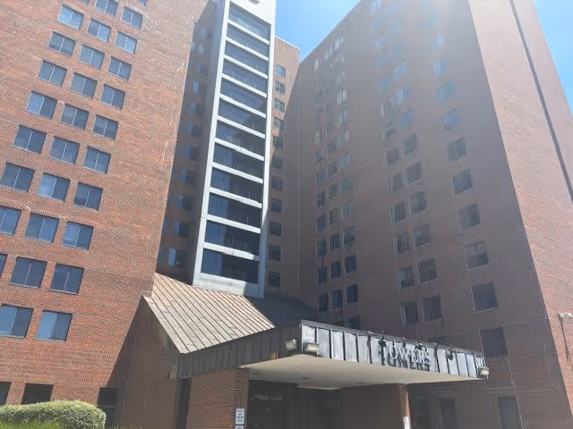 Exterior view of a tall brick senior living facility building with multiple windows and a covered entrance labeled 'Towers'. The sky is clear and sunny.