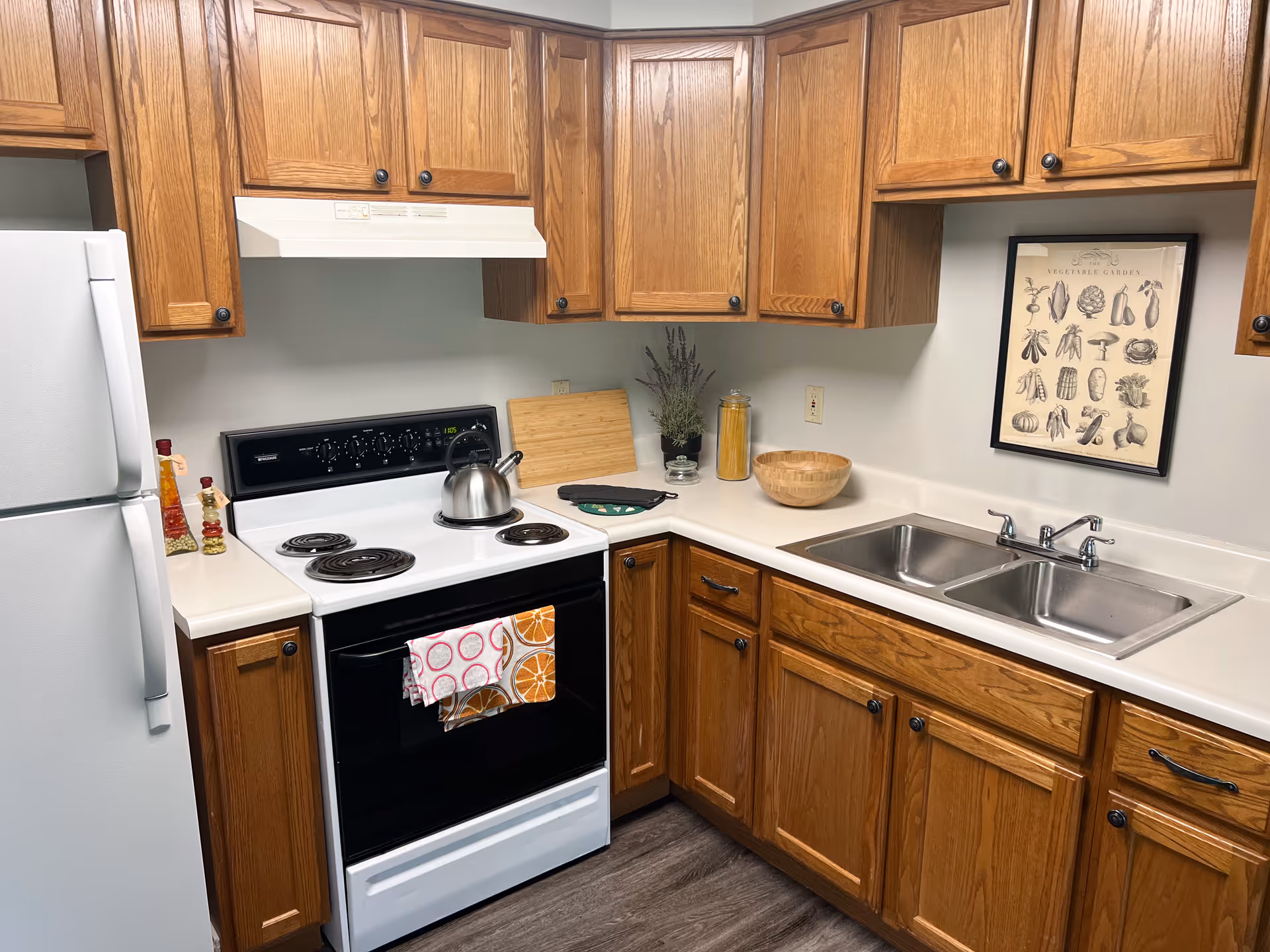 A kitchen with wooden cabinets, a white refrigerator, a white electric stove with a kettle on it, and a double stainless steel sink. The countertop has a cutting board, a potted plant, a jar of pasta, a wooden bowl, and two oven mitts. A framed vegetable garden print hangs on the wall above the sink.