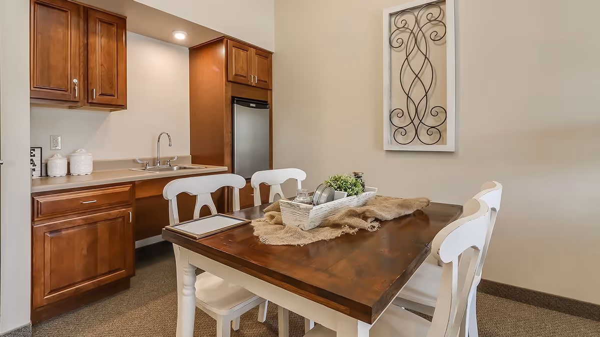 A small dining area with a wooden table and four white chairs. On the table, there is a decorative centerpiece with a small plant and other items on a burlap cloth. Behind the table, there is a kitchenette with wooden cabinets, a sink, and a small refrigerator. A decorative wrought iron wall hanging is mounted on the beige wall.