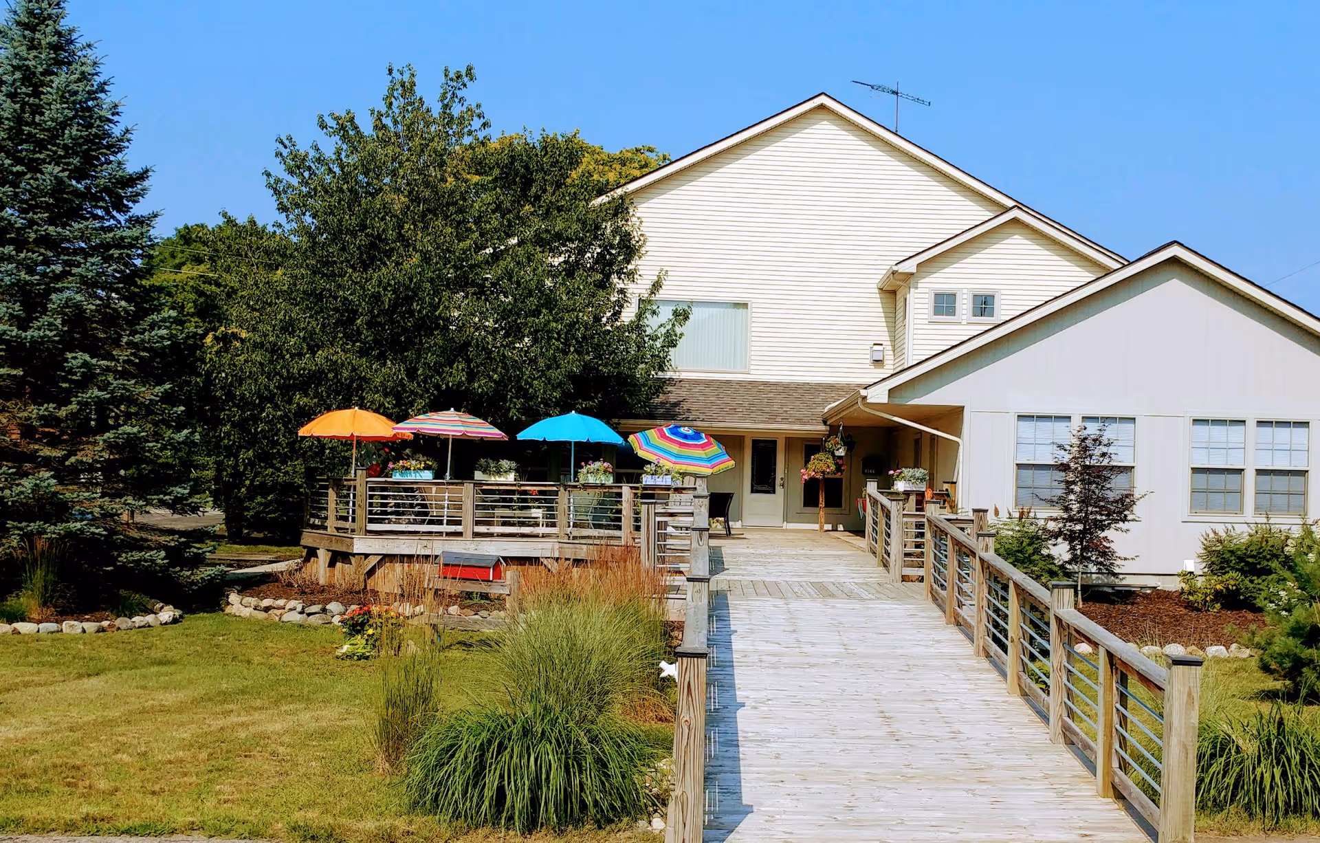 Exterior view of a senior living facility with a wooden ramp leading to the entrance. The building is light-colored with multiple windows. There is a deck area with colorful umbrellas and potted plants. Surrounding the building are green trees, shrubs, and a well-maintained lawn under a clear blue sky.
