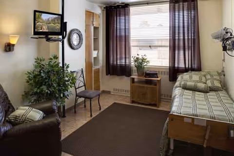 A small, cozy room with a single bed on the right side covered with green and beige striped bedding. There is a brown leather armchair with a matching pillow on the left side, a metal chair next to a tall green plant, and a small wooden cabinet under a window with brown curtains. A wall-mounted TV and a round clock are visible on the left wall. The room has a brown carpet and light-colored walls.