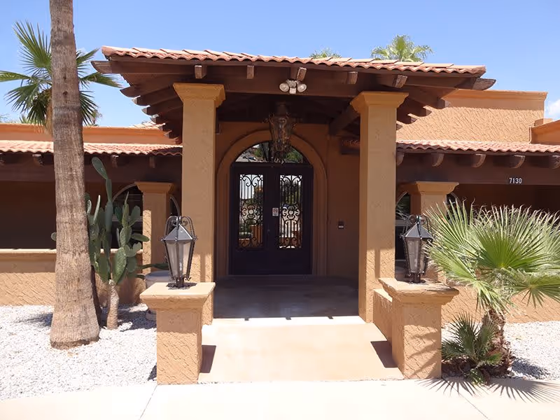 Entrance to a building with a southwestern architectural style featuring tan stucco walls, a tiled roof, two columns supporting a covered porch, decorative lanterns on the columns, and desert landscaping with palm trees and cacti.