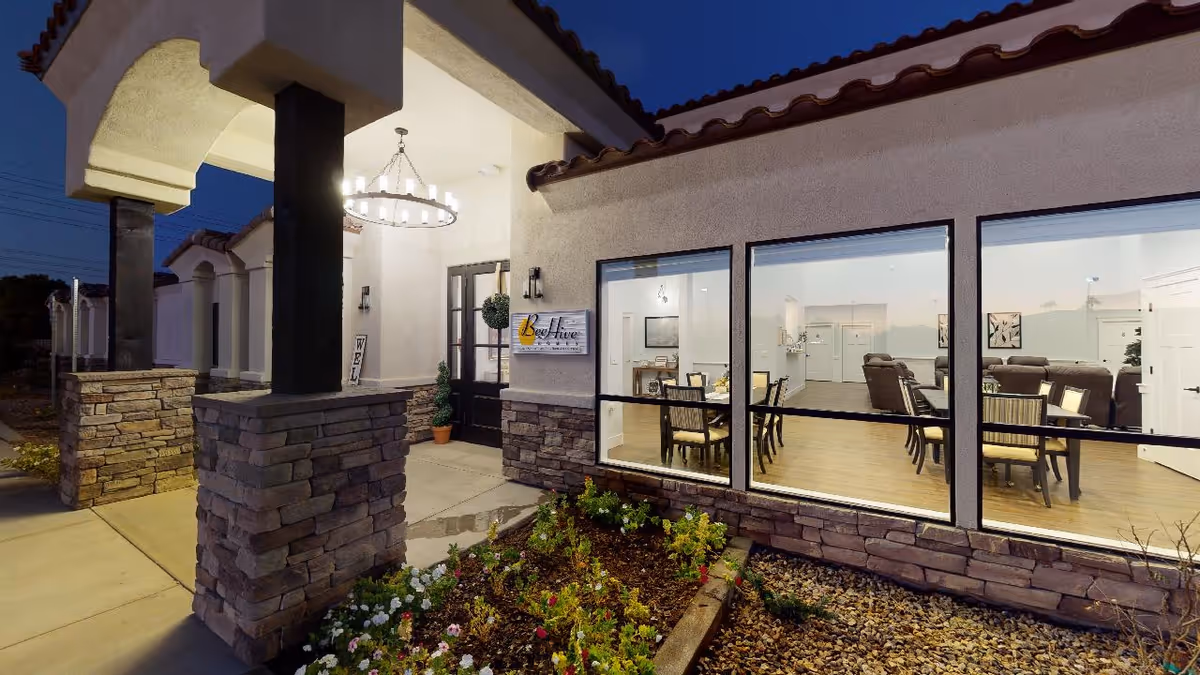 Exterior view of BeeHive Homes Assisted Living facility at dusk, showing the entrance with stone pillars, a hanging chandelier, and large windows revealing a well-lit interior dining and living area with tables, chairs, and sofas.