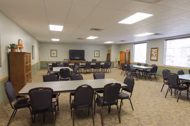 A spacious room with multiple tables and chairs arranged for group seating, featuring a large flat-screen TV mounted on the far wall, framed artwork, and windows allowing natural light to enter. The room has a carpeted floor and a drop ceiling with fluorescent lighting.