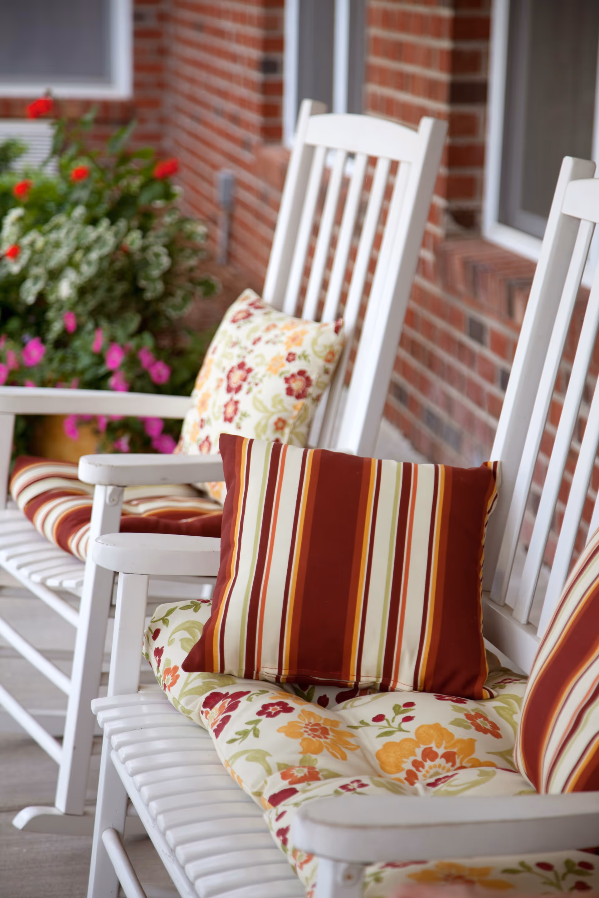 White wooden rocking chairs with floral and striped cushions on a porch with a brick wall and flowering plants in the background.