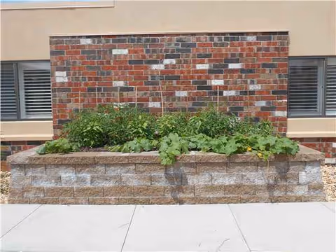 A brick building with a raised stone planter and green plants in front of a sidewalk.