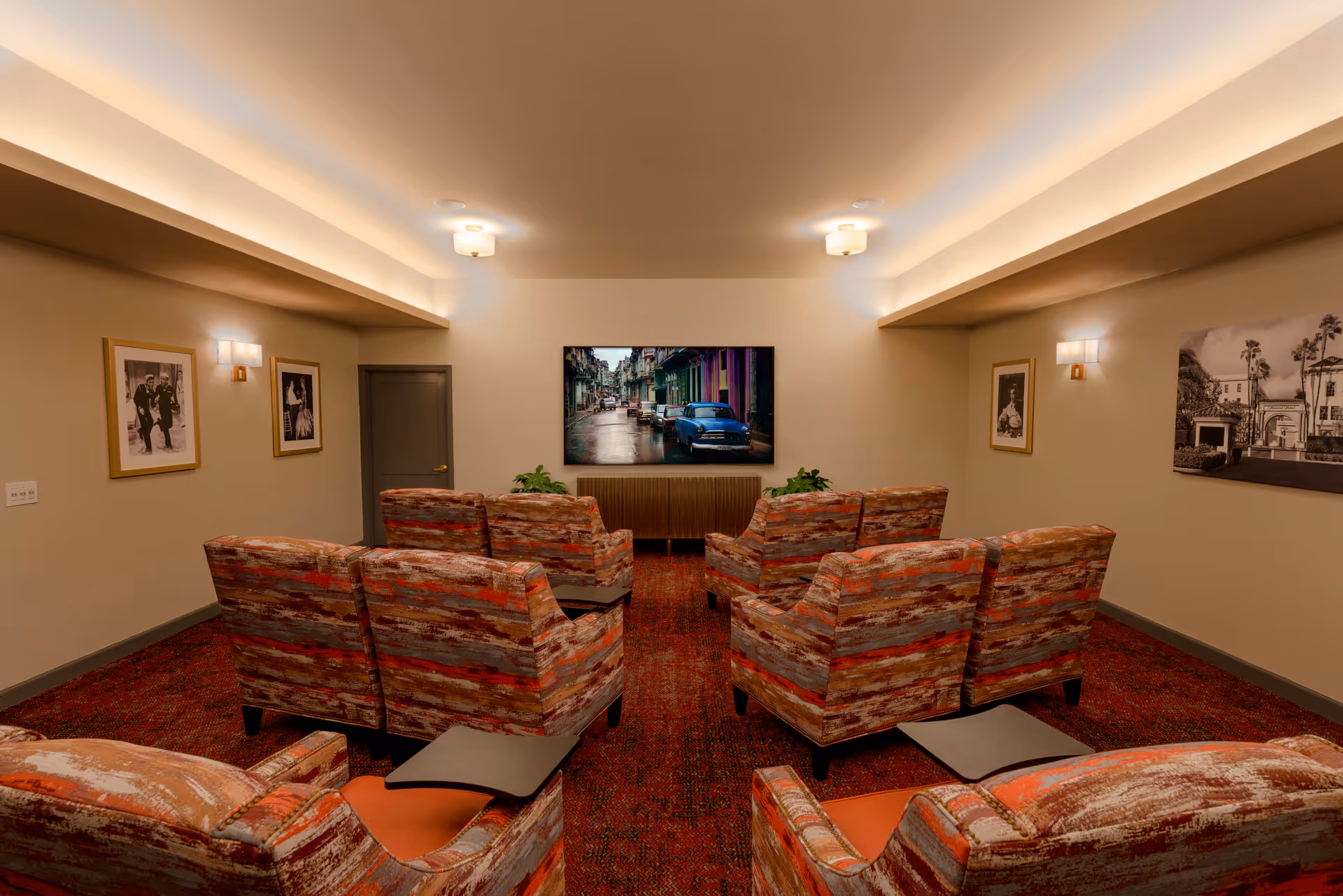 A cozy media room with multiple patterned armchairs arranged in rows facing a large flat-screen TV mounted on the wall. The room has warm lighting, framed black and white photographs on the walls, and a red patterned carpet.