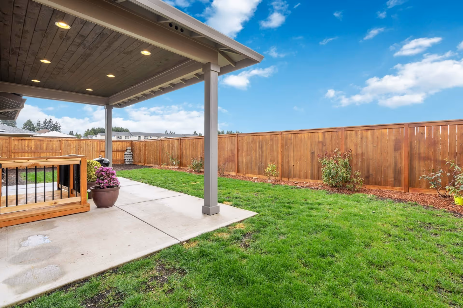 A backyard with a covered patio area featuring recessed lighting in the ceiling. The patio has a concrete floor and wooden railing on one side. The yard is grassy with a wooden fence enclosing the area and some small plants along the fence line. The sky is partly cloudy and blue.
