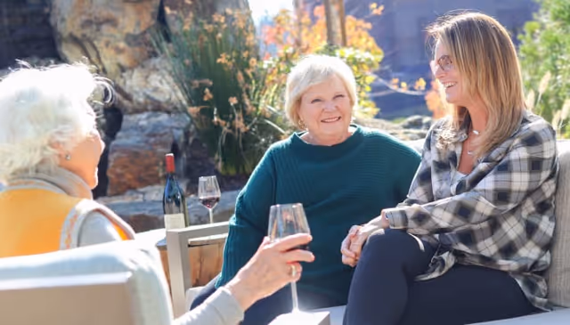 Three women sitting outdoors on patio furniture, enjoying conversation and holding glasses of red wine, with a rock waterfall and plants in the background.