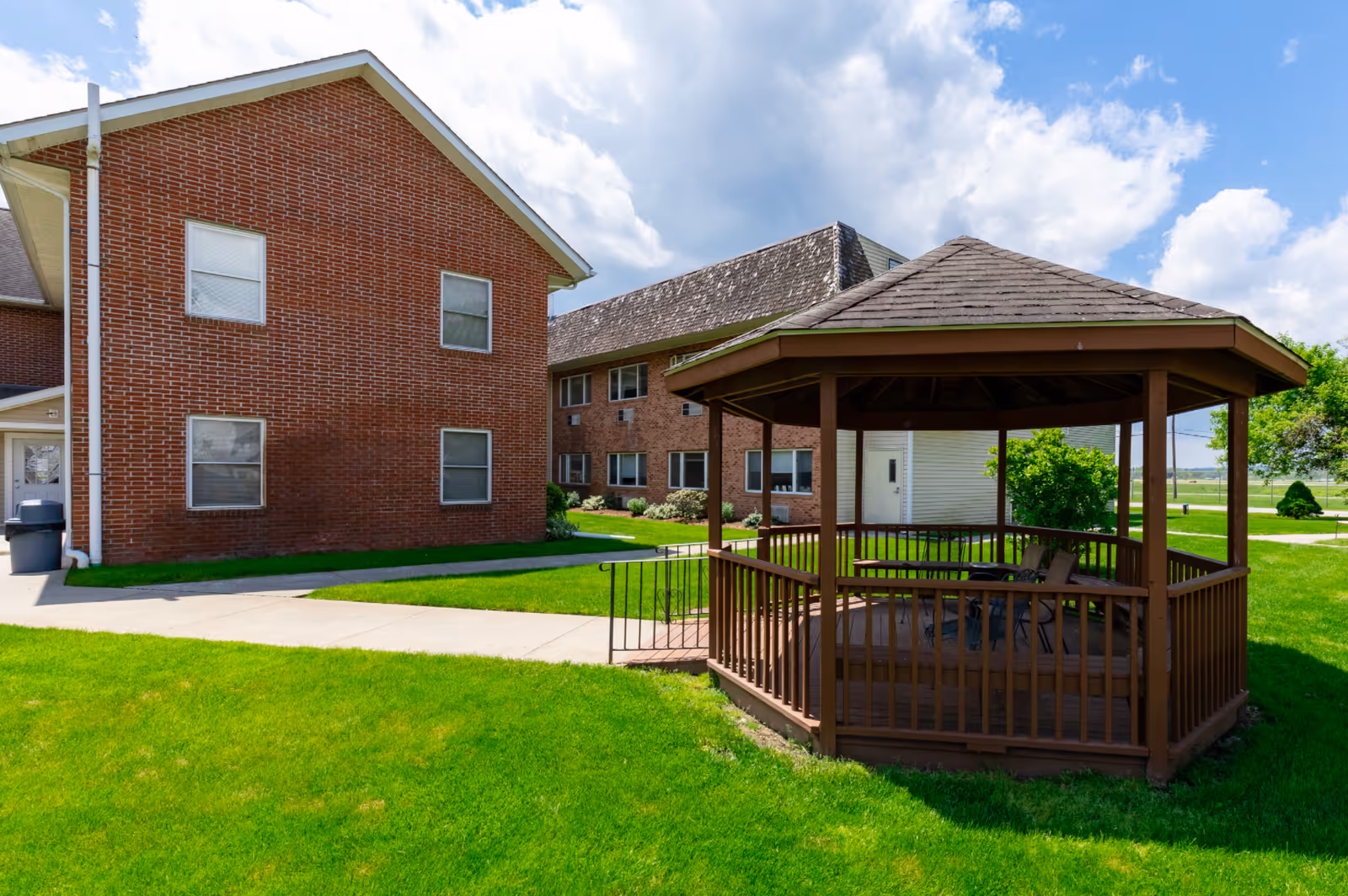Outdoor view of Avalon Assisted Living facility showing a brick building with multiple windows and a wooden gazebo with chairs on a well-maintained green lawn under a partly cloudy sky.