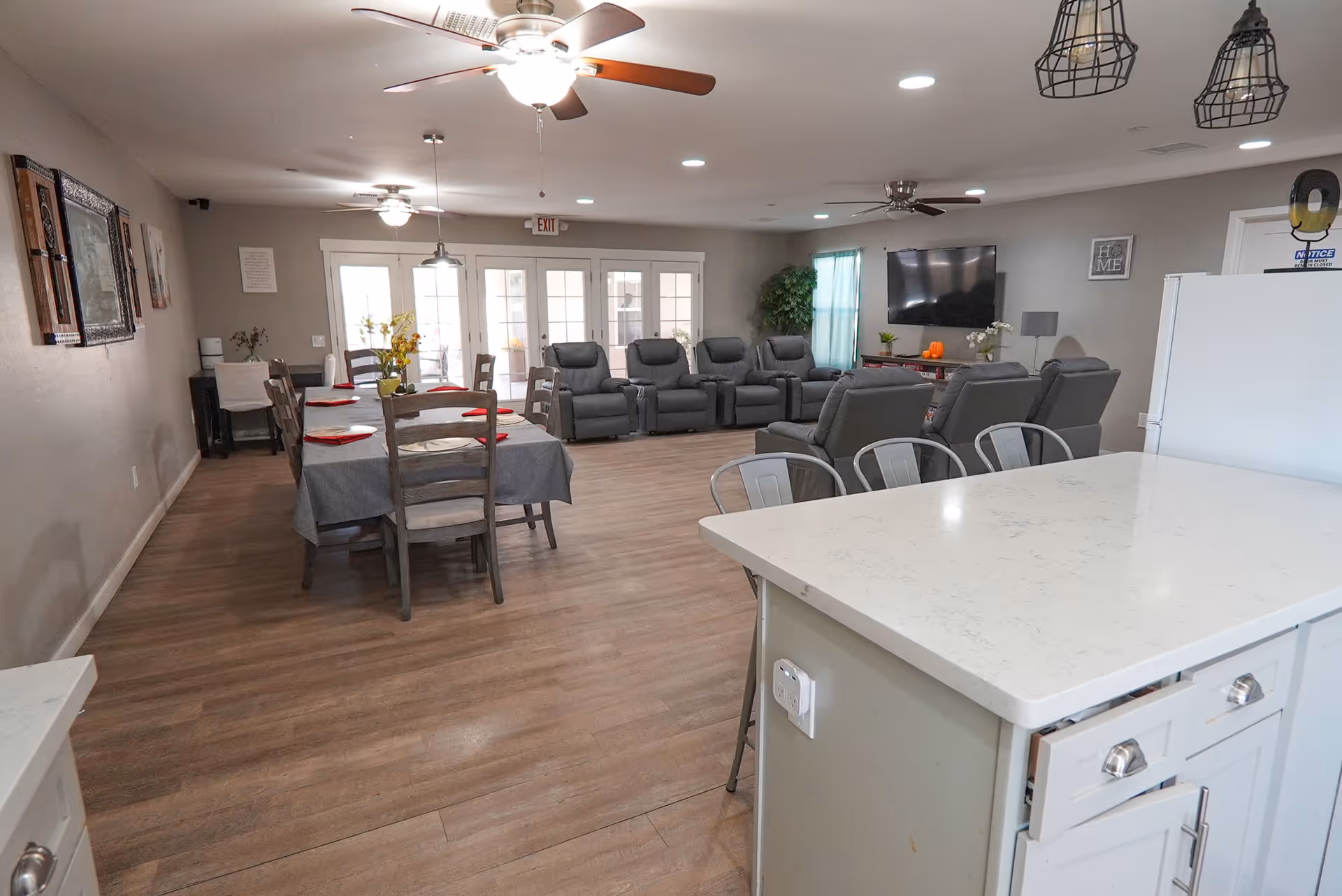 A spacious assisted living common area featuring a dining table with chairs and place settings, a row of comfortable recliner chairs facing a wall-mounted flat screen TV, ceiling fans, and a kitchen island with bar stools in the foreground. The room has wood flooring, neutral-colored walls, and large windows with curtains allowing natural light.