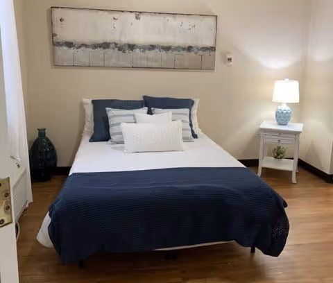 A neatly made bed with white sheets, multiple pillows in white and blue, and a dark blue blanket at the foot. The bed is positioned against a beige wall with a large abstract painting above it. To the right of the bed is a white nightstand with a blue and white lamp and a small plant underneath. The floor is wooden, and there is a large dark green vase on the left side near the wall.