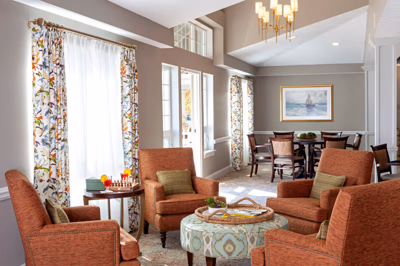 Bright communal living area with four orange armchairs around a patterned ottoman, a side table with a chess set, and a dining table in the background.