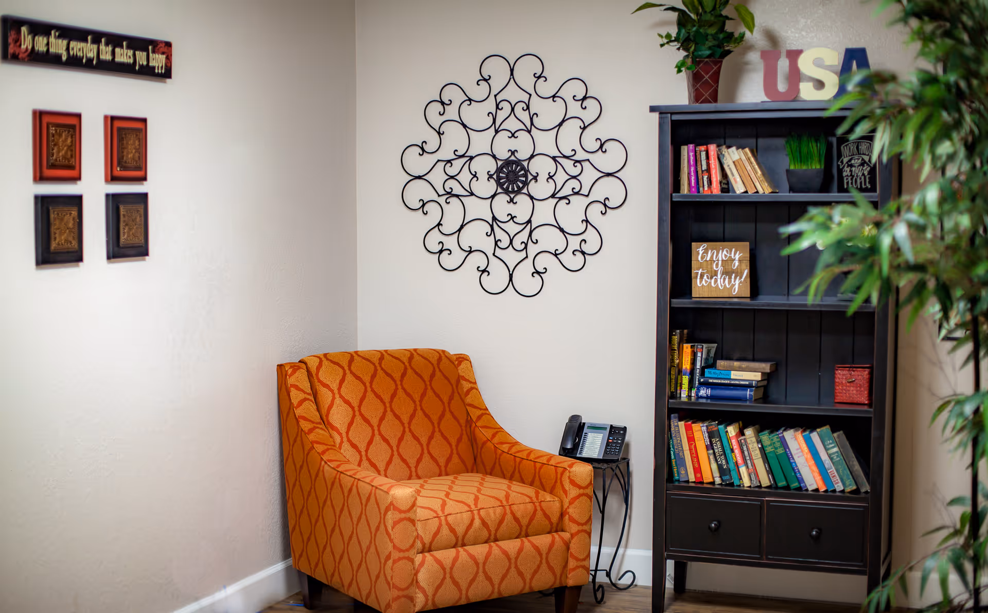 A cozy corner in a living room featuring an orange patterned armchair next to a small side table with a telephone. On the wall above the chair is a decorative black wrought iron wall art. To the right is a dark wooden bookshelf filled with books, a small plant, and decorative signs including one that says 'Enjoy today!' and wooden letters spelling 'USA'. There are also framed art pieces and a motivational sign on the adjacent wall.