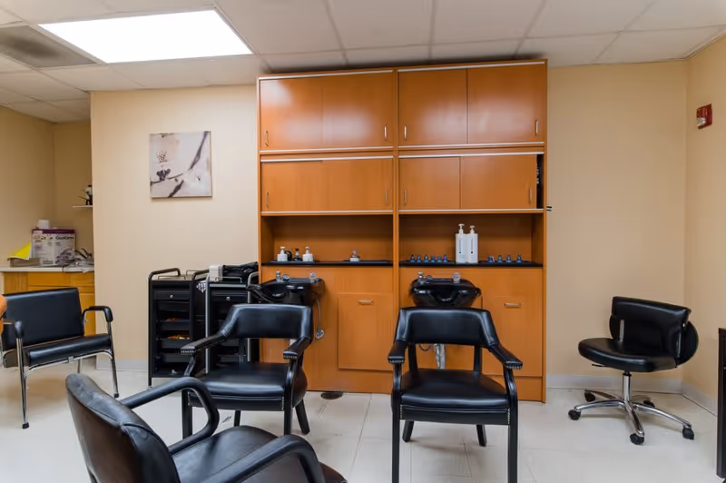 Interior view of a small salon area with four black salon chairs arranged around two black hair washing sinks built into wooden cabinetry. The cabinetry has multiple storage compartments and shelves with bottles of hair products. The room has beige walls, a tiled floor, and a ceiling with fluorescent lighting. There is a small piece of wall art on the left wall and a rolling cart with supplies.