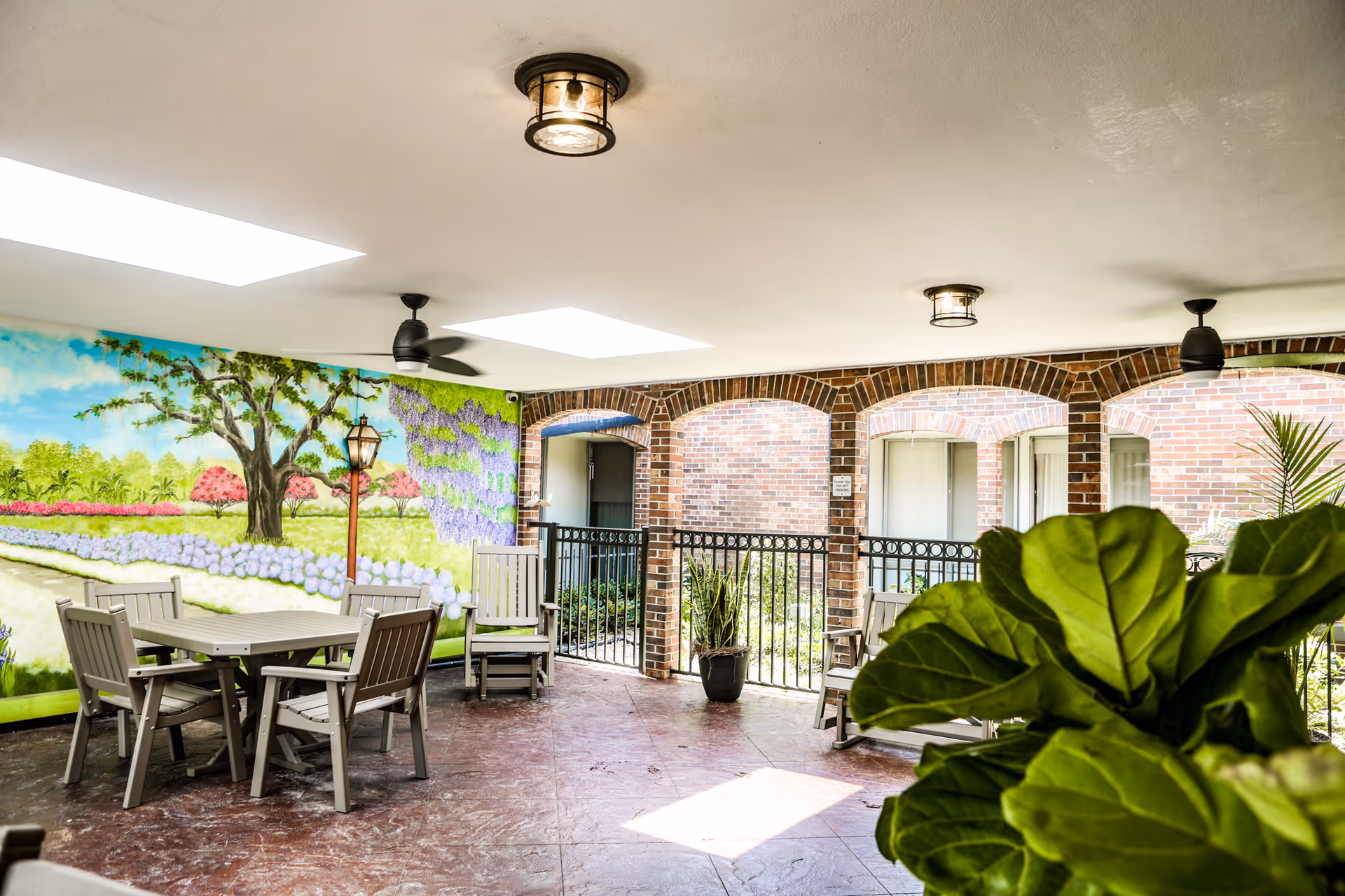 Covered outdoor patio area with tables and chairs, a mural-painted wall, brick arches and potted plants.