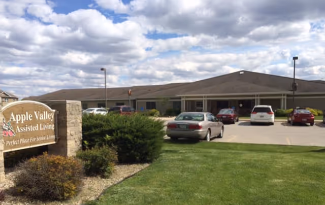 Exterior view of Apple Valley Assisted Living facility showing a single-story building with a parking lot in front, several parked cars, a green lawn, bushes, and a sign that reads 'Apple Valley Assisted Living Perfect Place For Senior Living' under a partly cloudy sky.