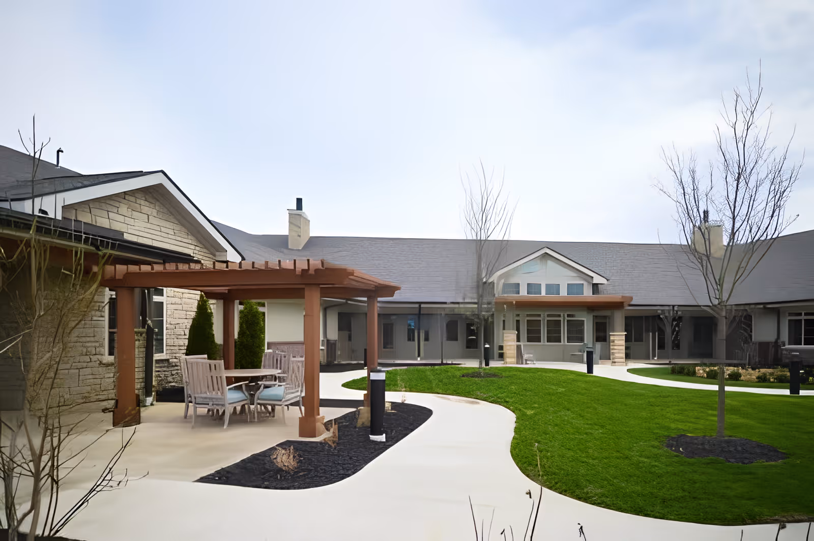 Courtyard of a senior living facility with a wooden pergola and outdoor dining set, curved walkways, green lawn, and surrounding single-story building.