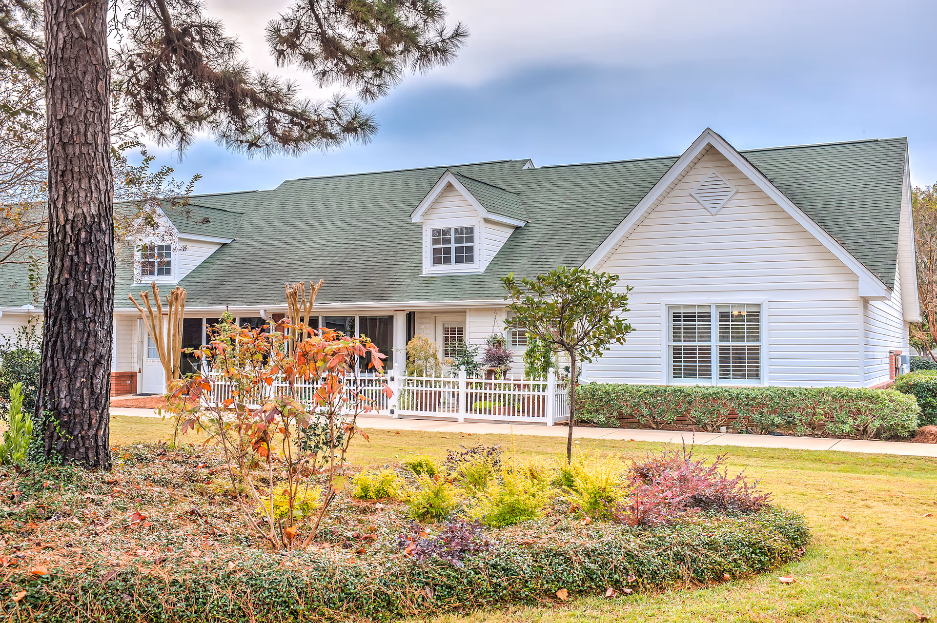 Exterior view of a single-story building with white siding and a green roof, surrounded by a well-maintained garden with various shrubs, small trees, and a white picket fence in front of the entrance.