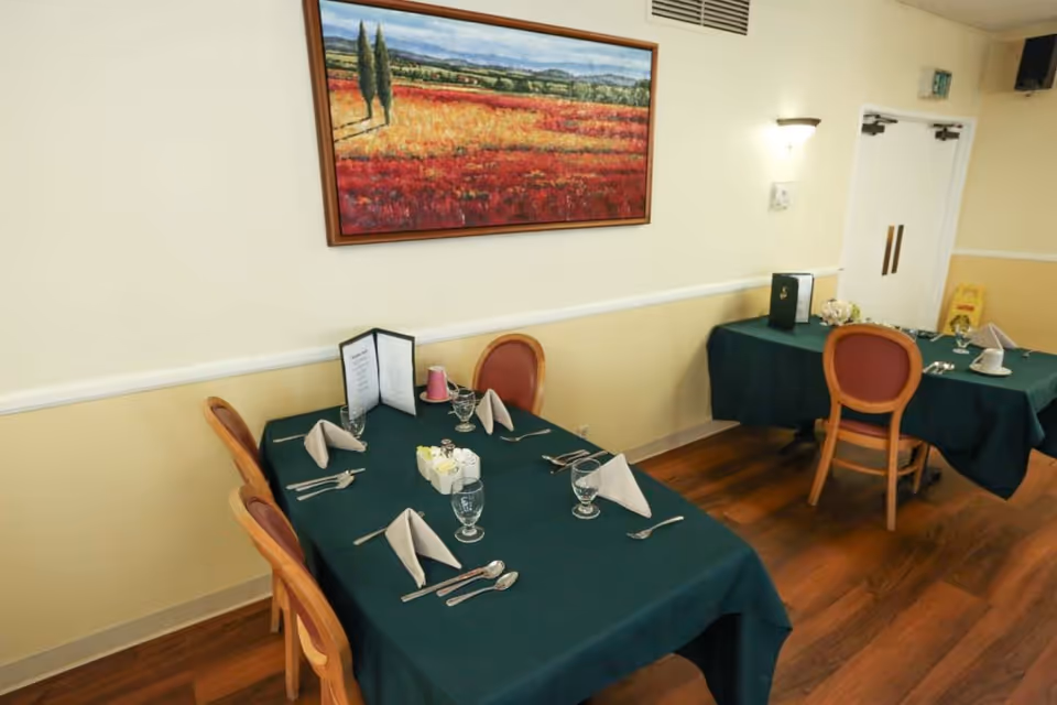 Dining area with two tables covered in dark green tablecloths, each set with folded napkins, glassware, utensils, and menus. The room has wooden flooring, cream-colored walls with a white chair rail, and a landscape painting hanging on the wall. There are wooden chairs with red cushioned seats around the tables.