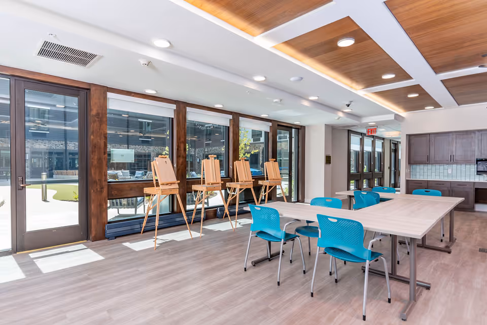 Bright interior activity room with large windows, tables and turquoise chairs and wooden easels by glass doors.