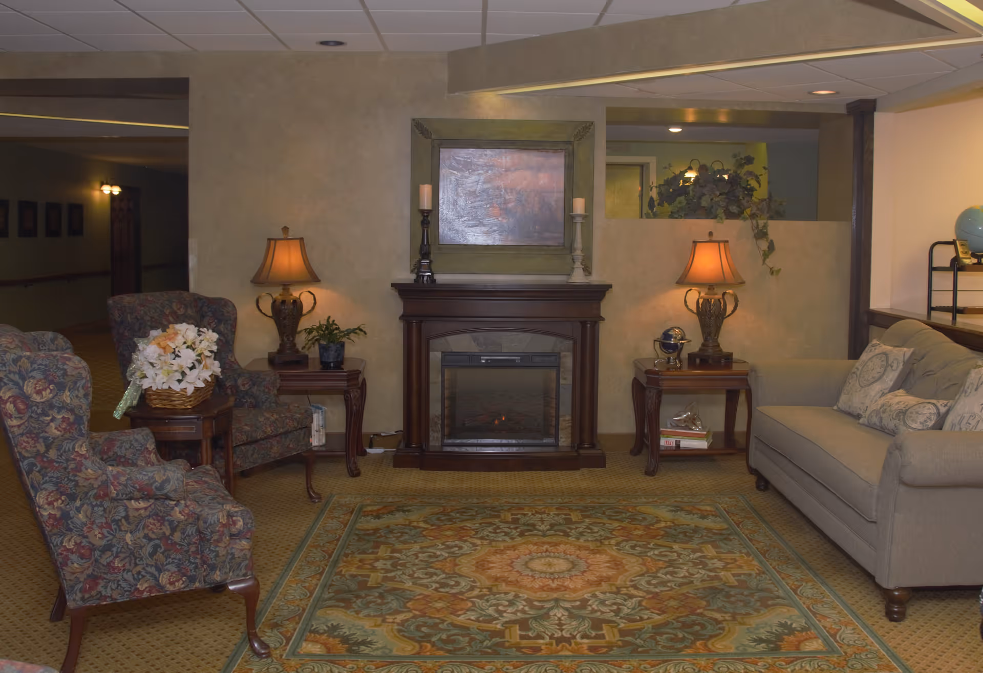 A cozy living room area with a patterned armchair and a beige sofa with decorative pillows. There is a wooden electric fireplace in the center with a framed painting above it and two table lamps on side tables flanking the fireplace. A floral arrangement is placed on a small table next to the armchair. The room has a patterned carpet and warm lighting.