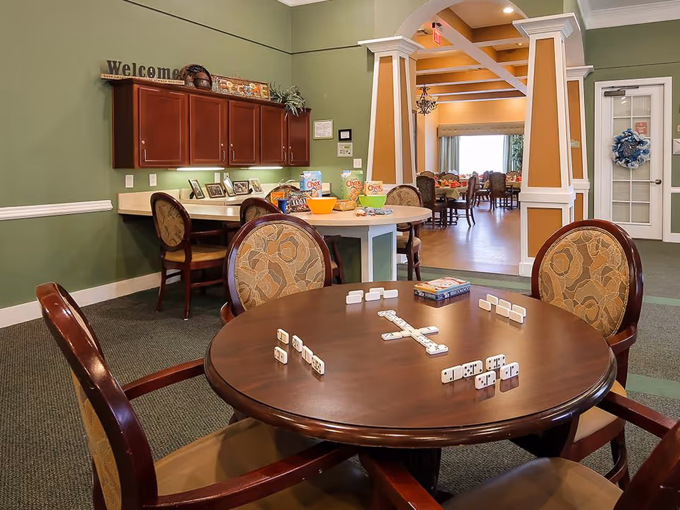 Activity/common room with a round table set for dominoes, chairs, a snack counter and cabinets with a 'Welcome' sign, and an archway leading to a dining area.