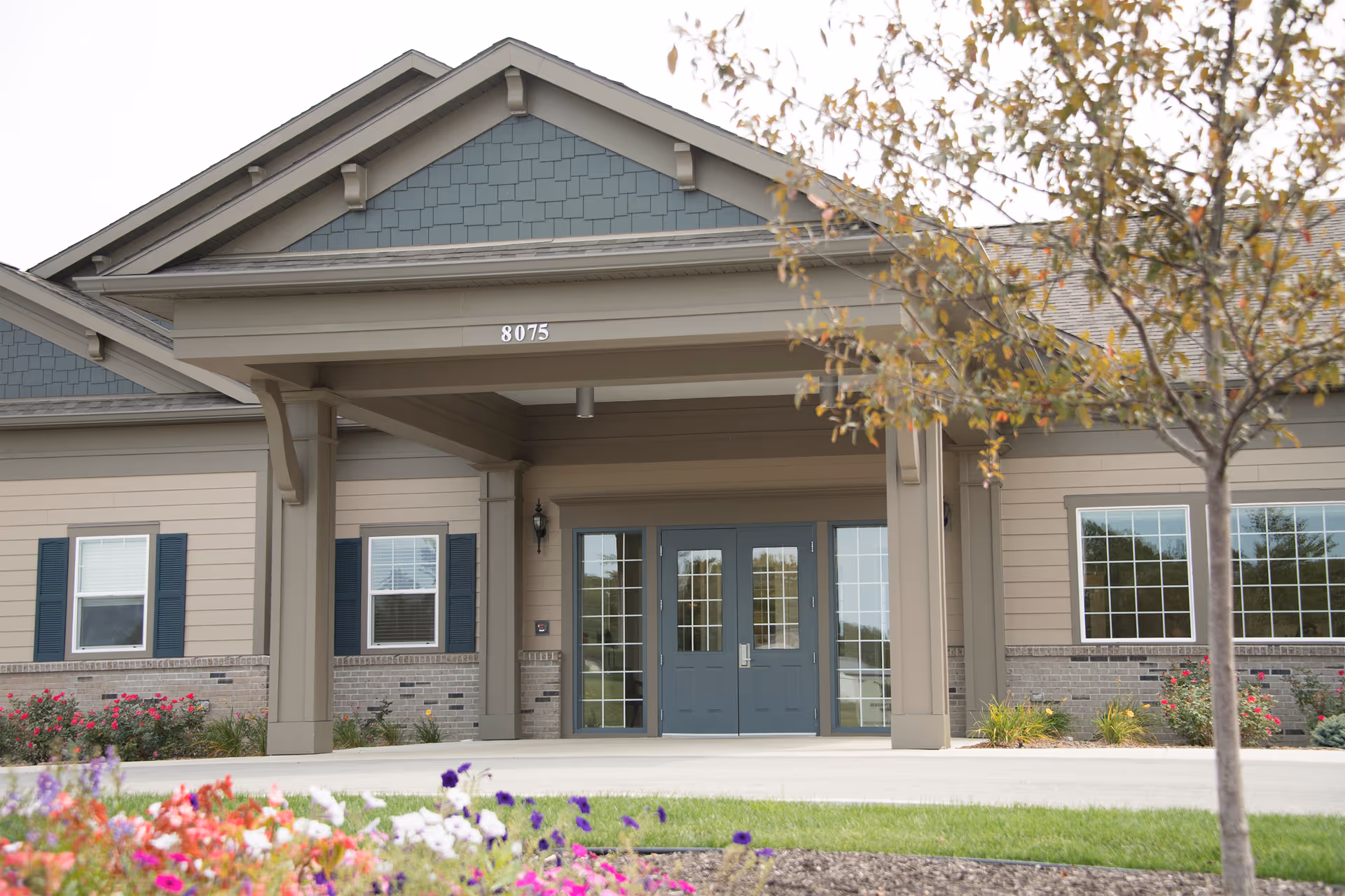 Front entrance of a single-story senior living building with double glass doors under a covered portico and landscaped flowerbeds.