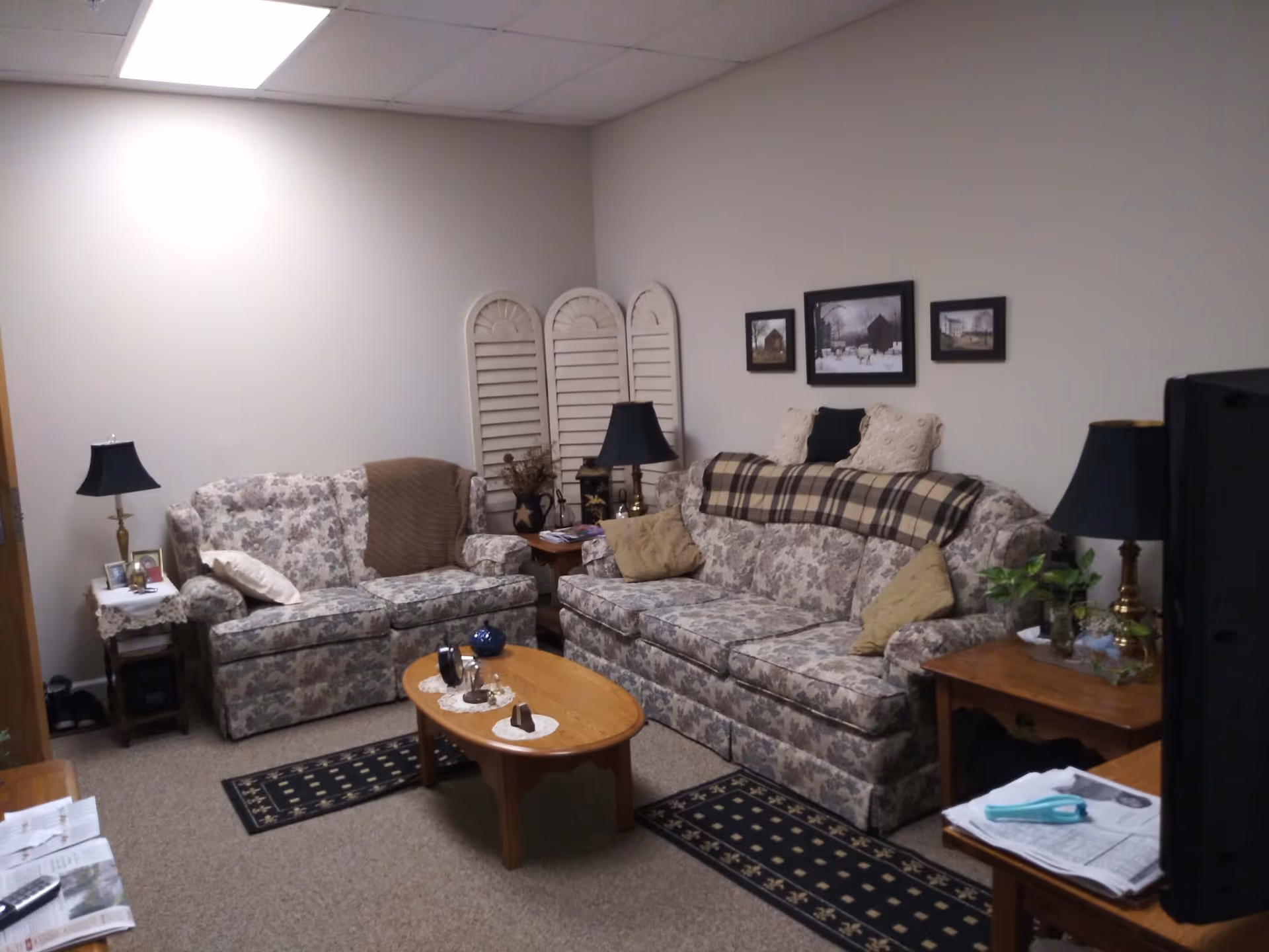 A cozy living room with two floral-patterned sofas, a wooden coffee table with decorative items, two side tables each with a lamp, and framed pictures on the wall. The room has beige walls and carpeted floor with small rugs.