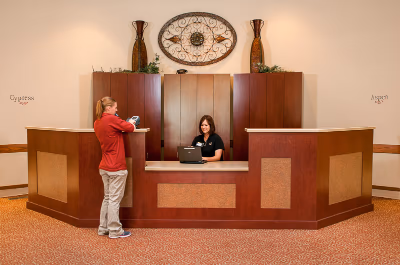 Reception desk in a senior living facility with a staff member at a laptop and a visitor standing at the counter.