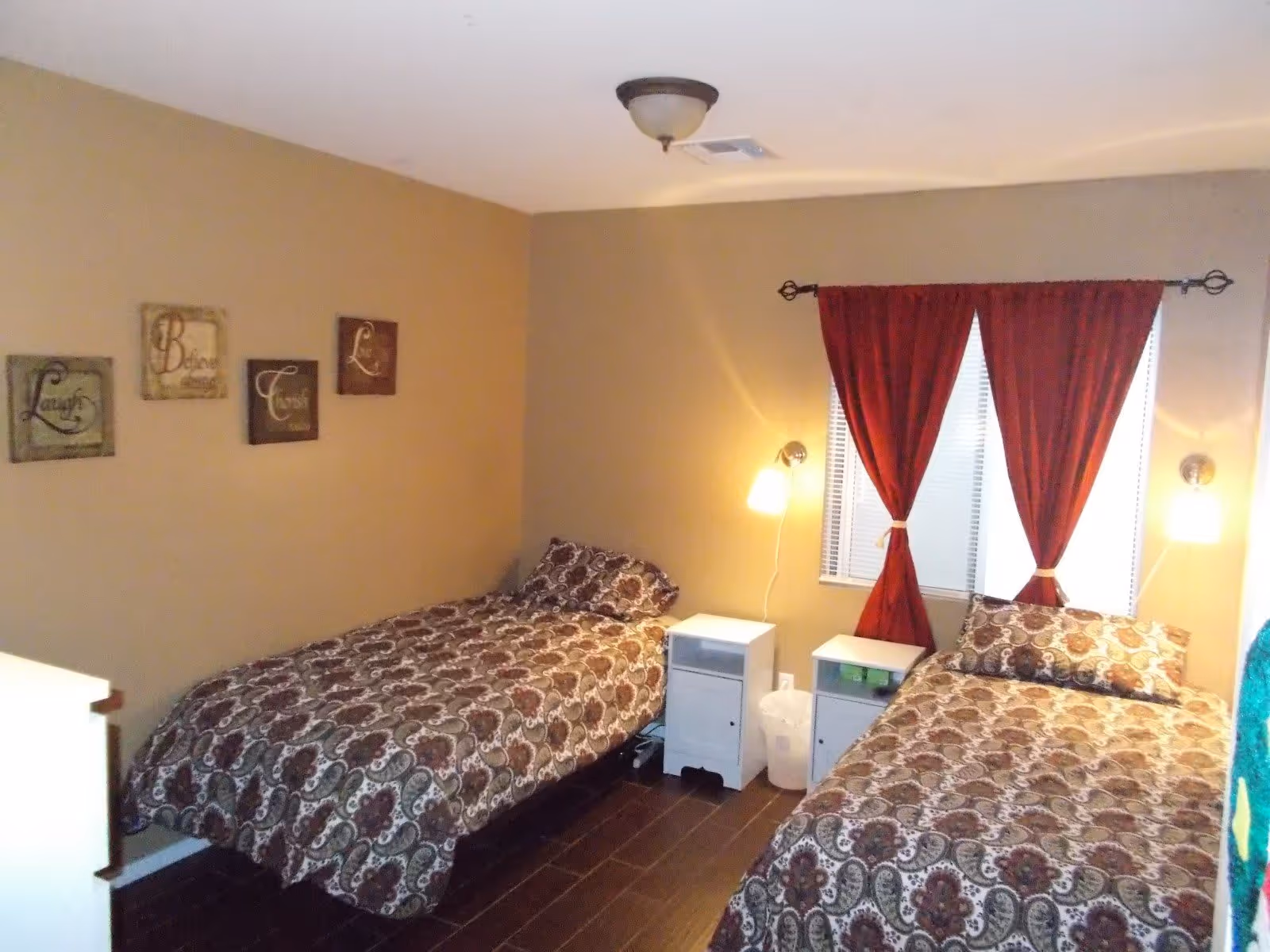 A bedroom with two single beds covered in matching patterned bedspreads. Each bed has a pillow and is positioned against adjacent walls. Between the beds are two small white nightstands with lamps mounted on the wall above them. A window with red curtains is centered on the wall behind the beds. The walls are painted beige and decorated with four framed inspirational word art pieces.