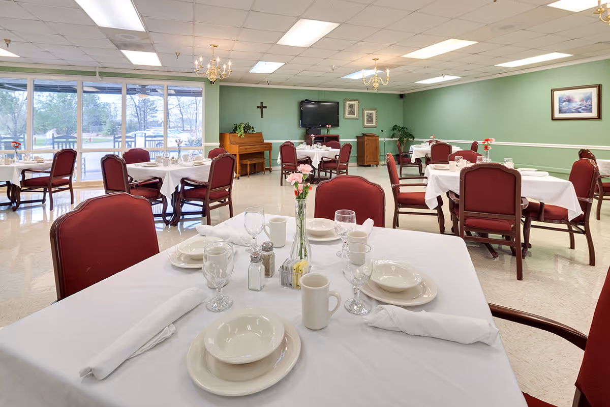 A dining room with multiple tables covered in white tablecloths, set with plates, bowls, glasses, mugs, and napkins. Each table has a small vase with flowers. The room has green walls, a piano, a TV mounted on the wall, framed pictures, and large windows letting in natural light. Red cushioned chairs surround the tables.