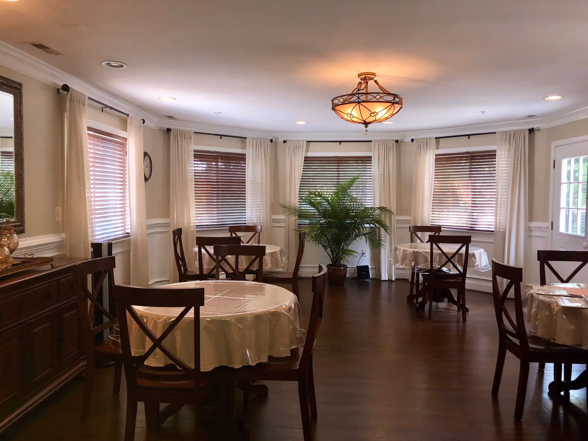 A dining room with several round tables covered with plastic tablecloths and surrounded by wooden chairs. The room has multiple windows with white curtains and wooden blinds, a large green potted plant in the corner, a wooden sideboard with decorative items, and a ceiling light fixture with a warm glow.