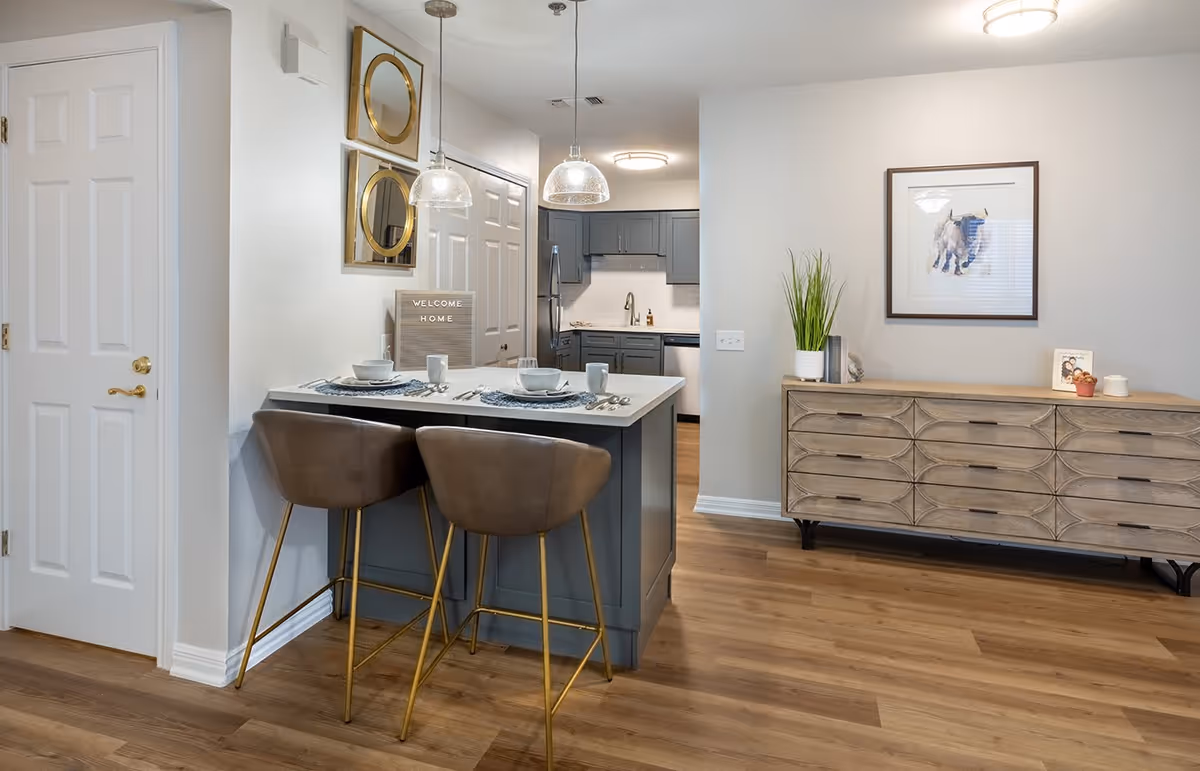 Modern kitchen and dining area with a small breakfast bar featuring two brown bar stools with gold legs. The kitchen has gray cabinets and stainless steel appliances. On the wall near the bar, there are two round mirrors and a sign that says 'WELCOME HOME'. To the right, there is a wooden sideboard with decorative items and a framed picture above it. The floor is light wood, and the walls are painted white.
