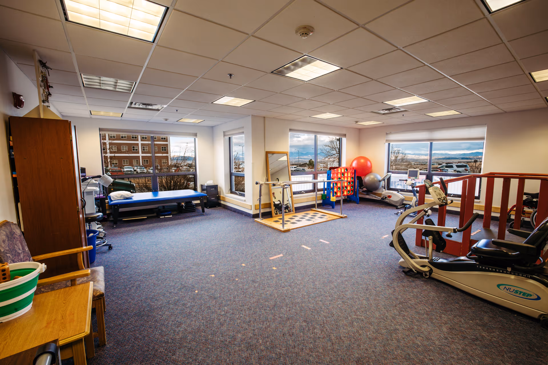 A spacious rehabilitation room with exercise equipment including a NuStep recumbent cross trainer, parallel bars for walking practice, a large red exercise ball, and a blue therapy table near the windows. The room has large windows letting in natural light and a carpeted floor.