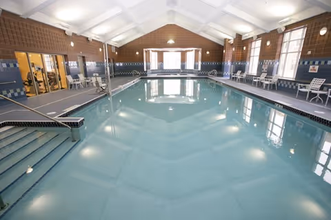 Indoor community swimming pool with a vaulted ceiling, lounge chairs and windows along the sides.