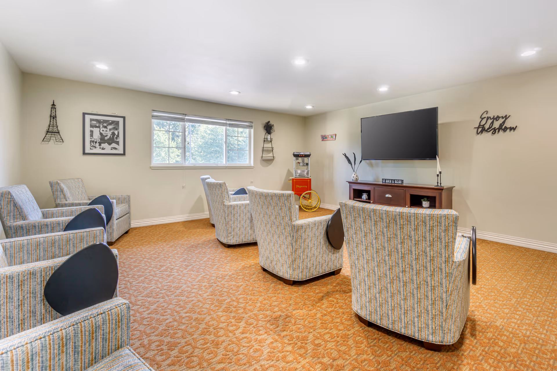 A cozy common room with several striped armchairs arranged in rows facing a large flat-screen TV mounted on the wall. The room has beige walls, a window letting in natural light, a popcorn machine in the corner, and wall decorations including a small Eiffel Tower sculpture, a black and white photo, and a sign that says 'Enjoy the show'.