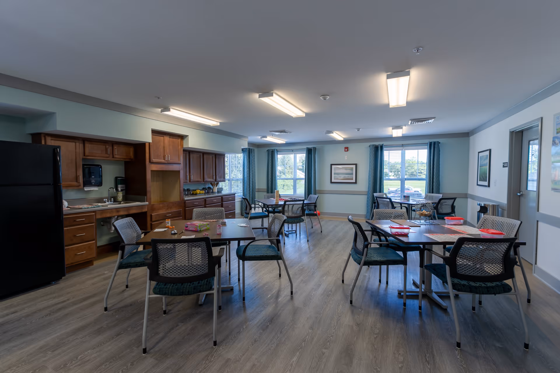 A well-lit communal dining area in a senior living facility featuring multiple tables and chairs arranged on a wood-style floor. The room includes a kitchenette with wooden cabinets, a black refrigerator, a sink, and countertop appliances. Large windows with blue curtains allow natural light to fill the space, and framed artwork hangs on the walls.