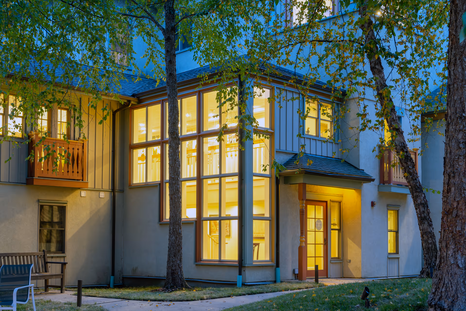 Exterior view of a senior living facility building at dusk with large illuminated windows, a small porch with a door, and trees with green leaves in the foreground. There is a bench and a chair on the left side near the building.