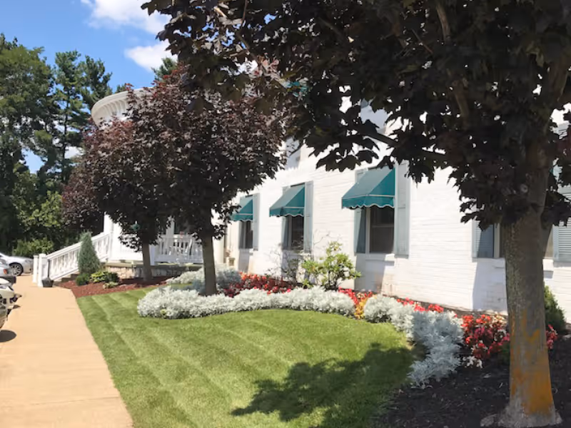 Front exterior of a white building with green window awnings, landscaped lawn, trees, and a sidewalk.