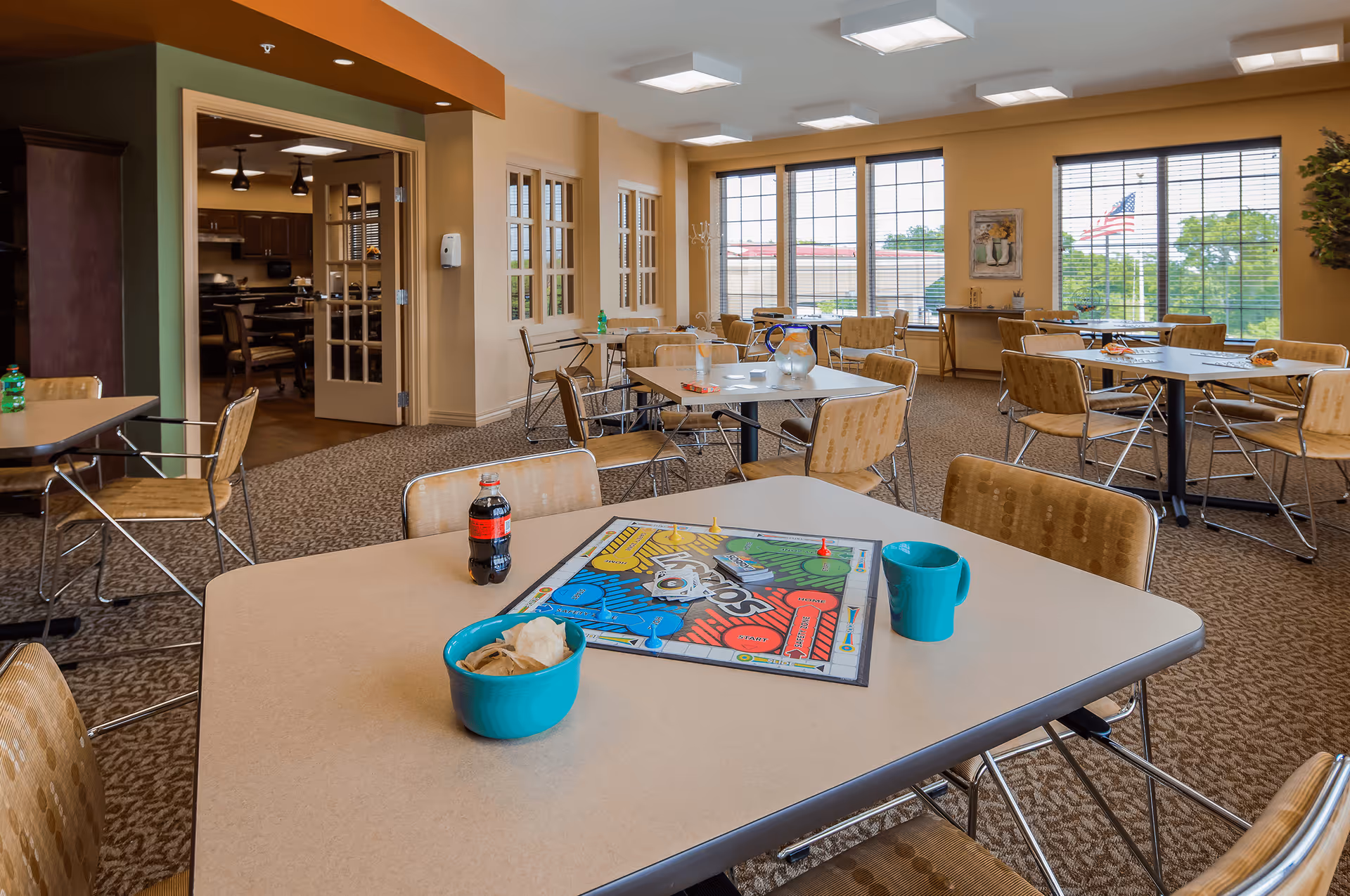 A bright and spacious common area with several tables and chairs arranged for group activities or dining. On the nearest table, there is a board game set up with game pieces, a bottle of soda, a blue bowl with chips, and a blue mug. Large windows with blinds allow natural light to fill the room, and an American flag is visible outside. The room has beige walls and carpeted flooring, with a doorway leading to another room that appears to be a kitchen or dining area.