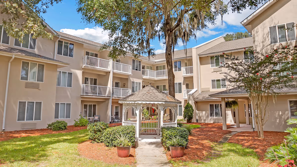 Outdoor courtyard area of a senior living facility with a white gazebo surrounded by green bushes and potted plants. The courtyard is enclosed by a three-story beige building with balconies and windows. Trees and a clear blue sky are visible in the background.