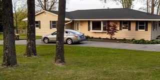 Single-story beige building with a dark roof, driveway, parked cars, and trees in foreground.