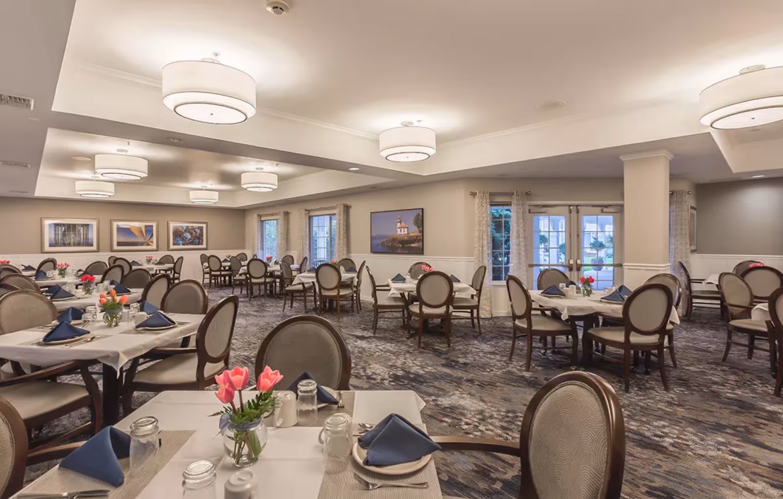 Spacious dining room with round tables set with napkins, flowers, and chairs under ceiling lights.