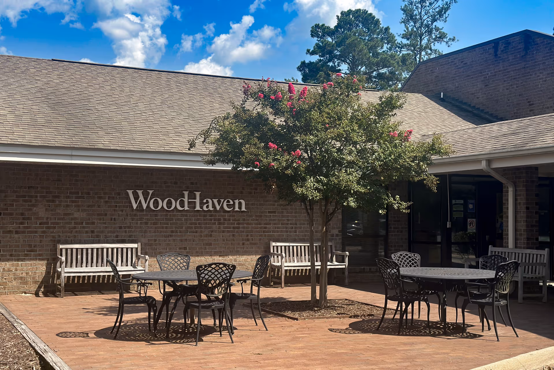 Outdoor patio area at WoodHaven Nursing Center with metal tables and chairs, wooden benches, a flowering tree in the center, and a brick building with the WoodHaven sign on the wall under a partly cloudy sky.