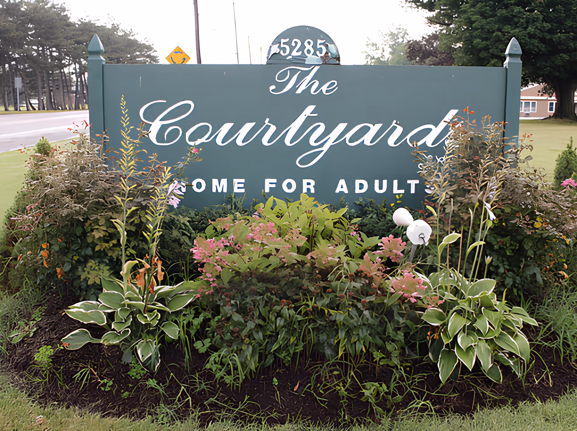 Green entrance sign for The Courtyards Adult Care with the address atop, surrounded by flowering plants and landscaping.