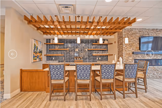Interior view of a retirement community lounge area featuring a wooden bar counter with six high chairs upholstered in blue floral fabric. Behind the bar is a decorative wall with shelves holding glassware and bottles, and a wooden ceiling trellis above. To the right, there is a stone wall with a mounted flat-screen TV and a fireplace below it.