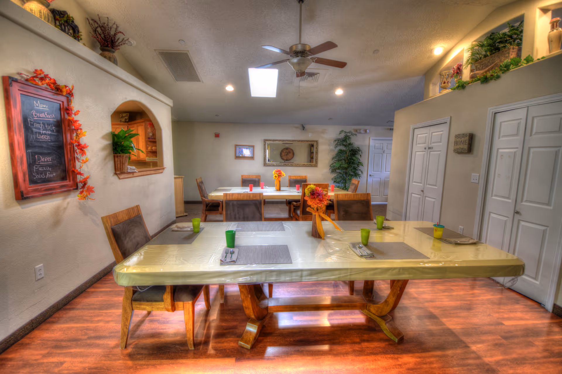 A dining room with two tables covered in plastic tablecloths, each set with placemats, utensils, and colorful cups. The room has wooden flooring, a ceiling fan, and recessed lighting. There is a chalkboard menu on the left wall decorated with autumn leaves, and a pass-through window to the kitchen. The back wall has a large mirror, framed pictures, and a tall green plant. Several doors and decorative plants are visible on the right side.