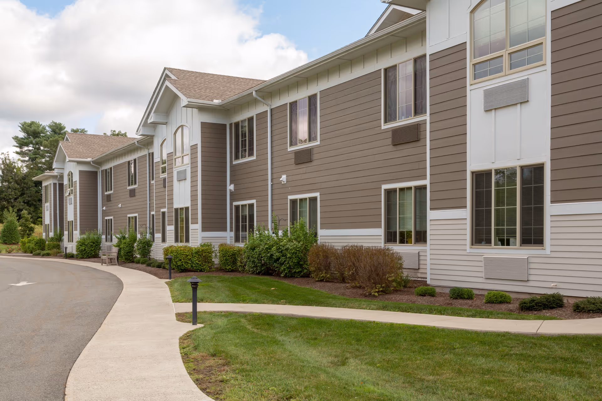 Exterior view of a two-story senior living facility building with beige and white siding, multiple windows, a curved sidewalk, and landscaped greenery including bushes and grass.